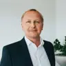 Mann mit blonden kurzen Haaren blickt leicht lächelnd in die Kamera, er trägt ein weißes Hemd und ein dunkles Jacket, rechts hinter ihm eine grüne Topfpflanze, die Wand dahinter ist hell